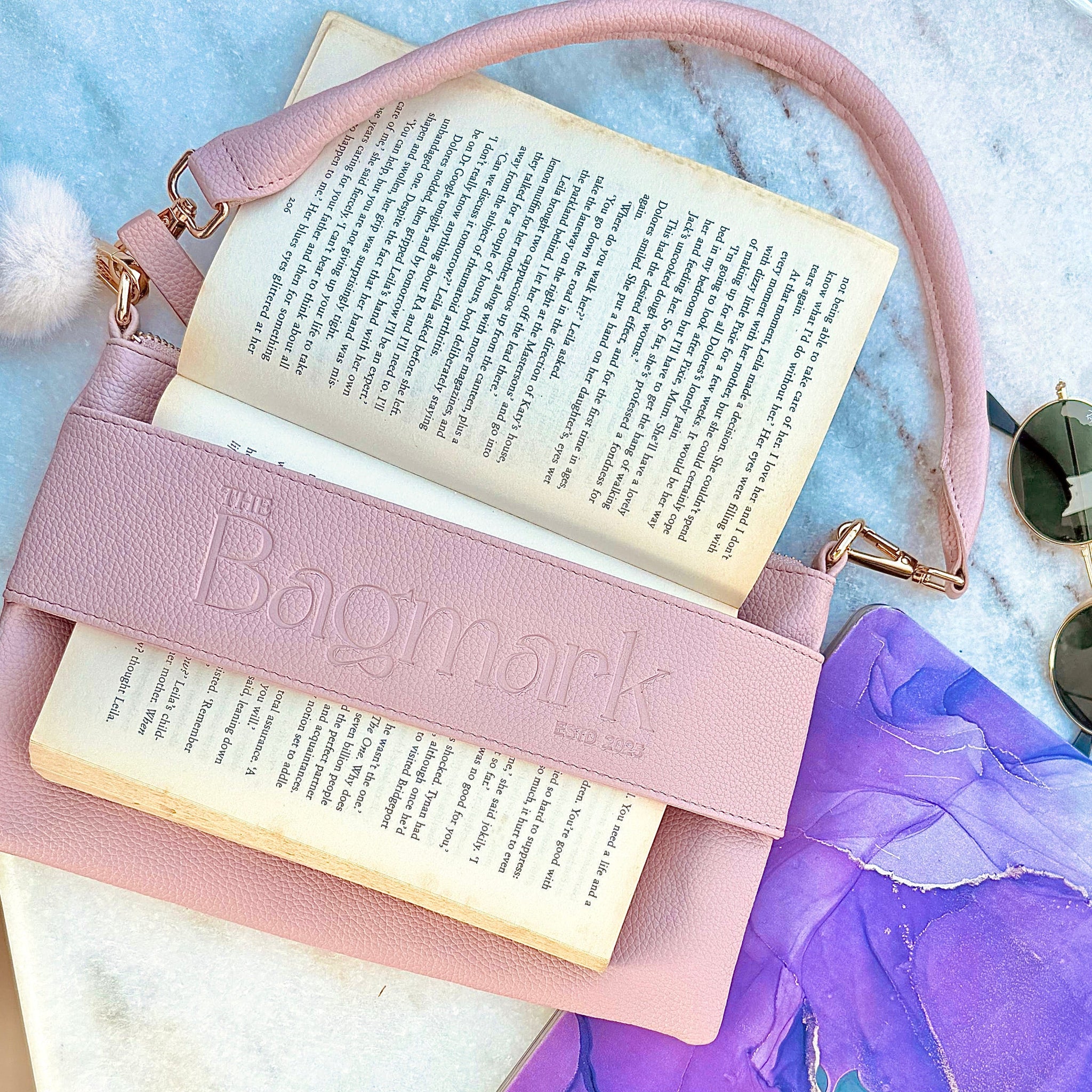 Pink handbag with 'The Bagmark' branding containing an open book on a textured surface.